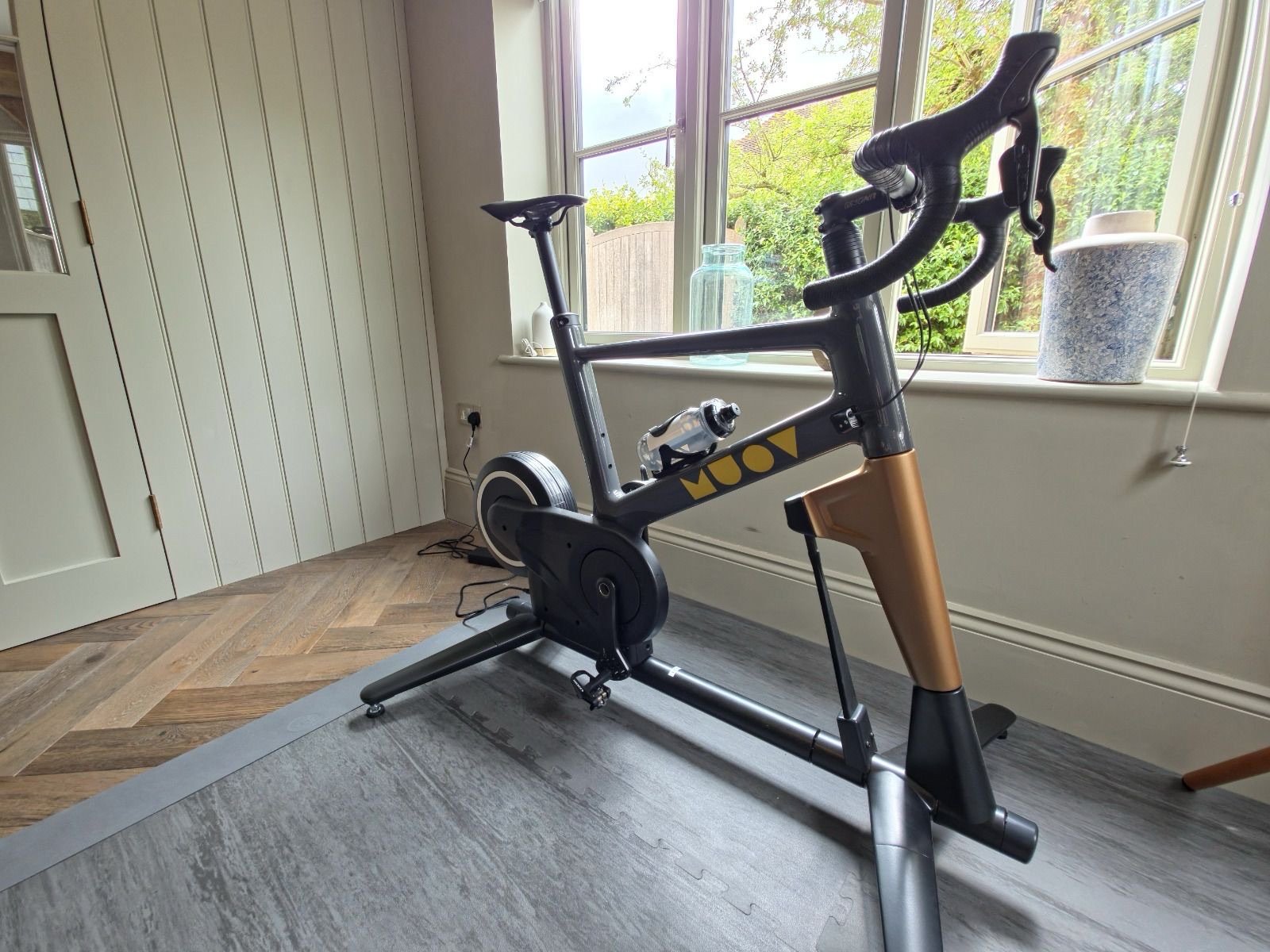 MUOV bike with a dark grey frame, gold lettering and gold fork, set up beside a window at home