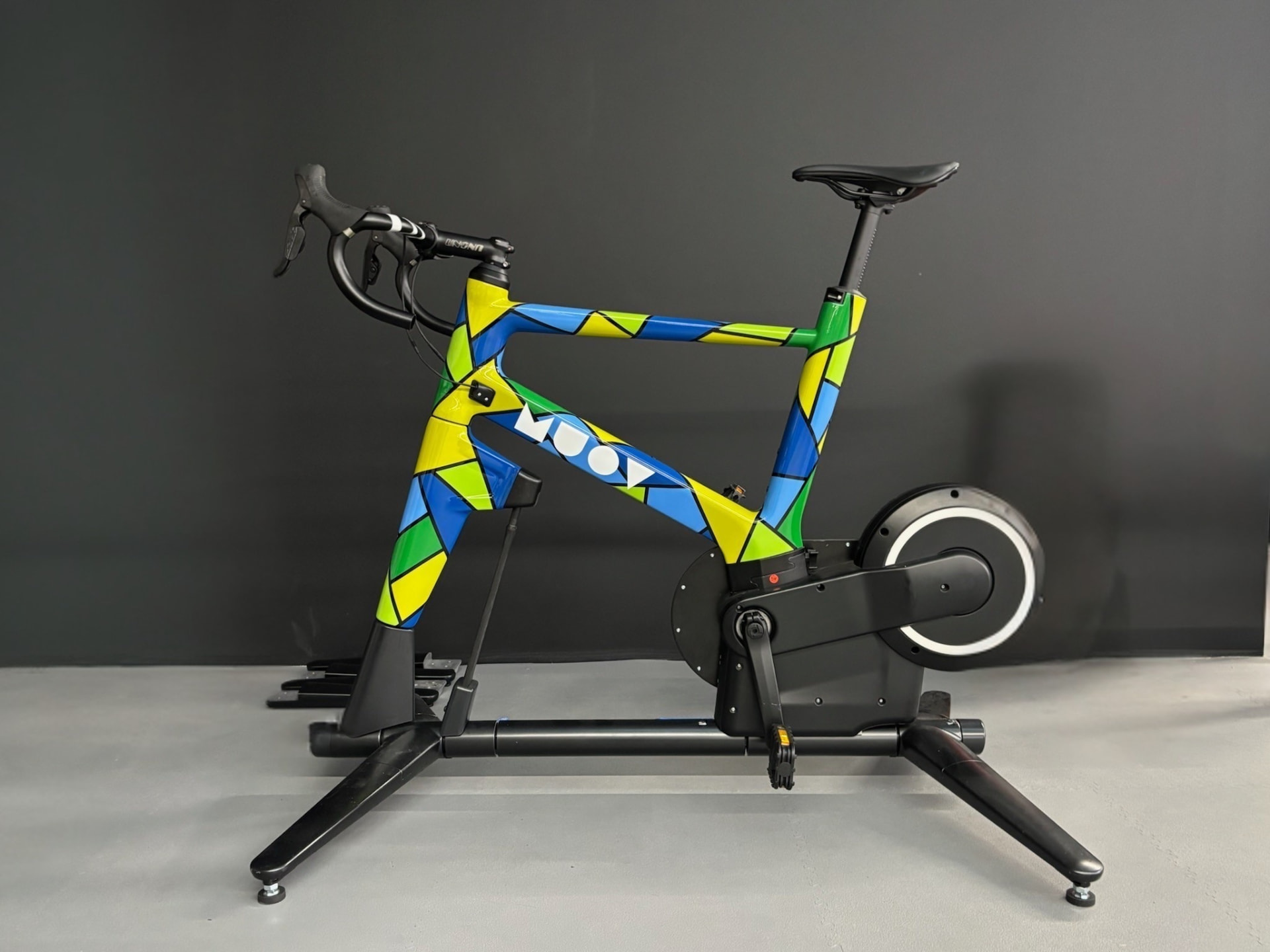 MUOV bike with a harlequin painted frame in blue, green and yellow facets, side-on against a dark grey wall