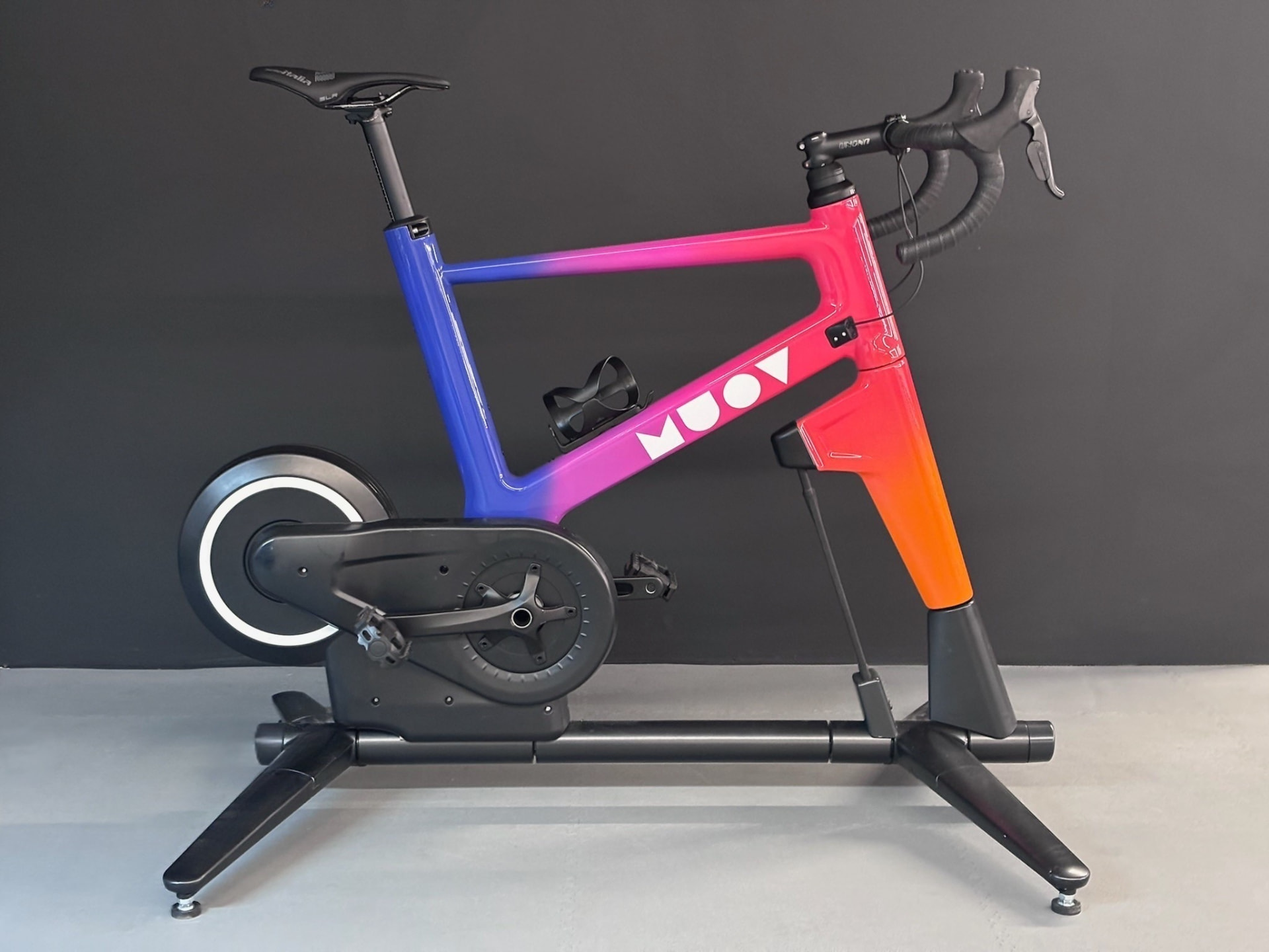 MUOV bike photographed side-on against a dark wall, its frame fading from blue through pink to orange with white MUOV lettering
