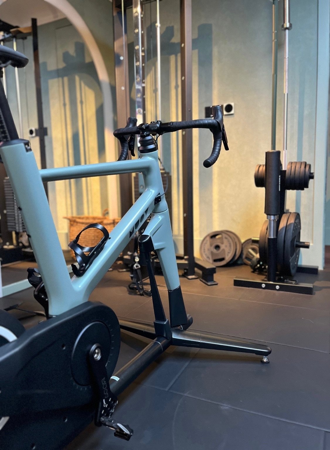 MUOV bike with a pale blue frame in a home training space with weights behind it